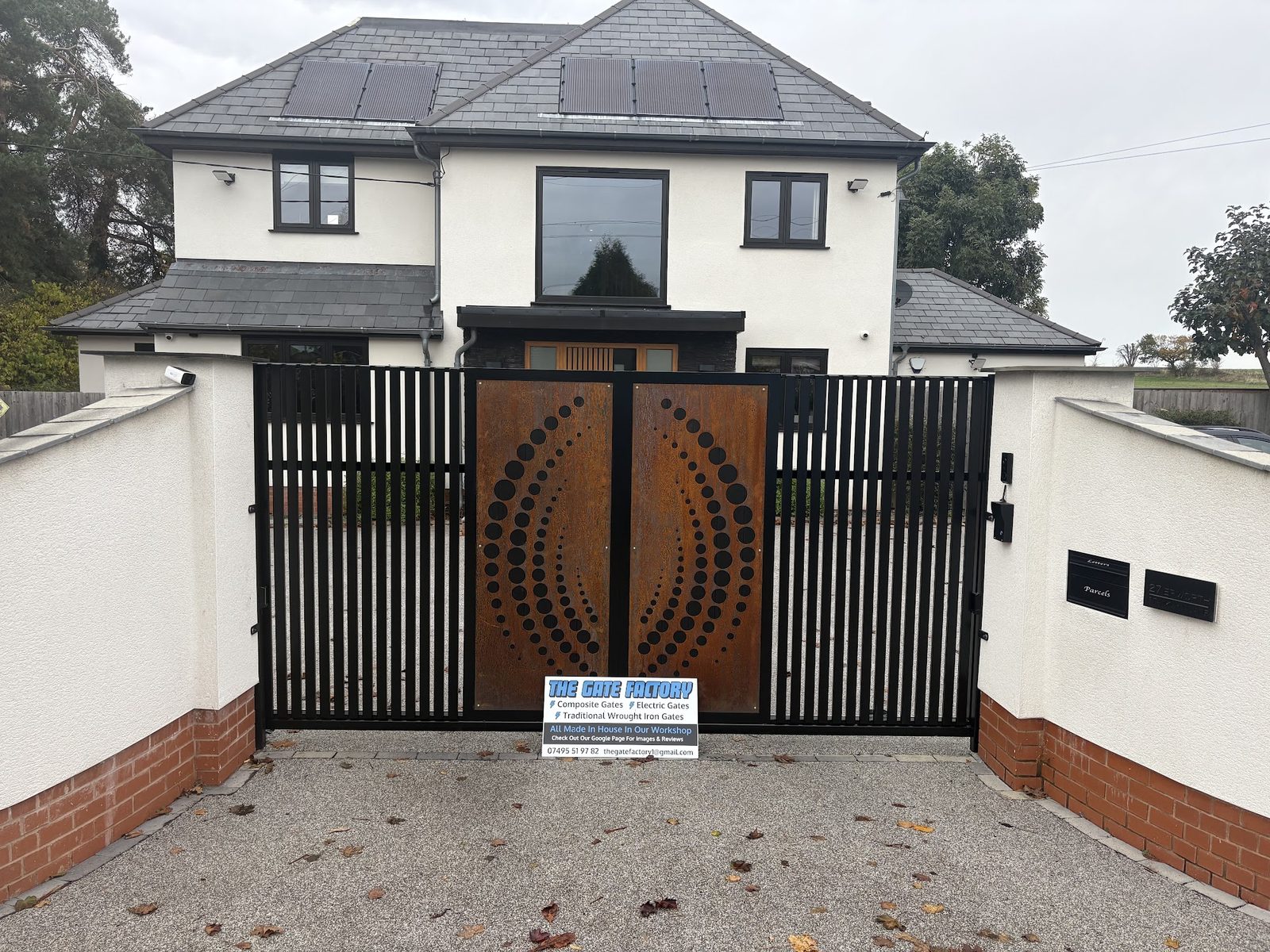 Black driveway gates with a corten steel sunburst panel, fitted to a rendered house